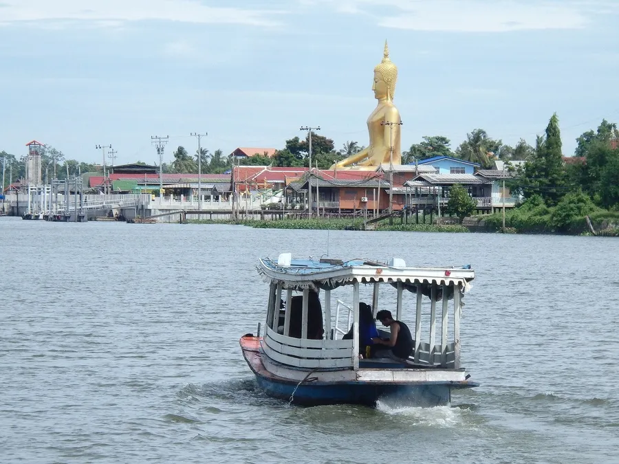 Barca cruzando el río Chao Phraya hacia Ko Kret, con casas sobre pilotes y un gran Buda dorado al fondo.